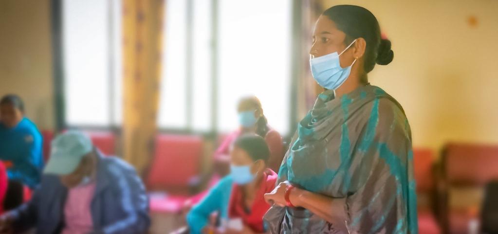 A woman is standing with her gray and blue shawl wrapped around her, and a mask covering half of her face.