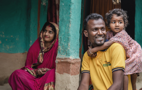 A woman wearing pink saree, red sindoor looks far ahead. Beside her is a man with a yellow t-shirt smiling and behind the man is an infant with a dress, hugging the man from behind.