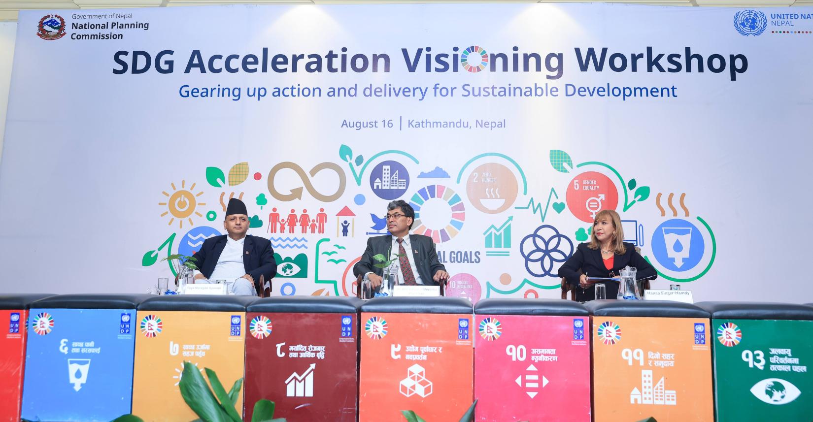 Dr. Toya Narayan Gyawali, Secretary, National Planning Commission (left), Dr. Min Bahadur Shrestha, Vice Chair, National Planning Commission (middle) and Ms. Hanaa Singer, UN Resident Coordinator, United Nations are sitting on stage with the SDG blocks lined in front of them and workshop banner behind them. The banners are filled with colorful SDG symbols