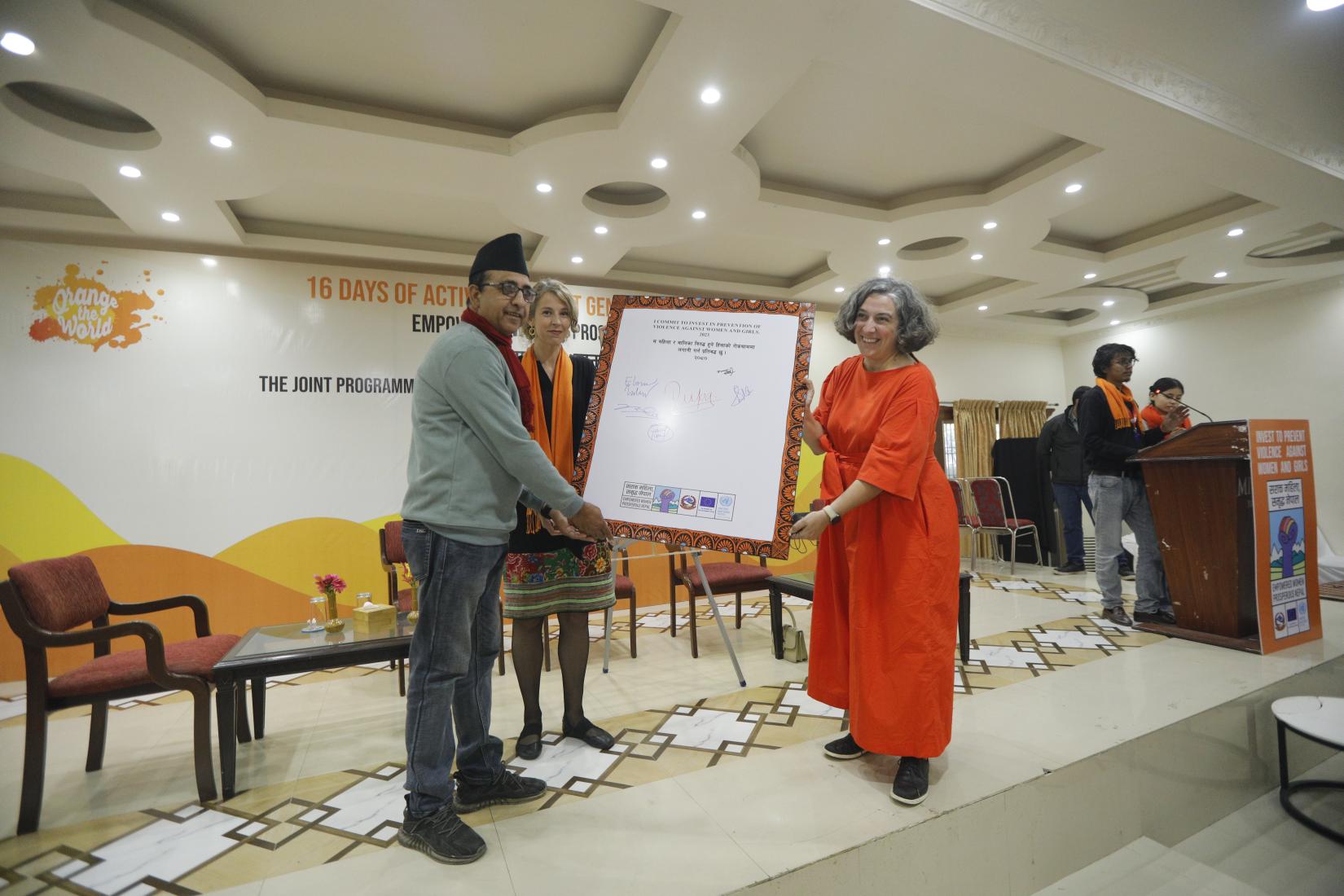 Two women and one man are standing on the stage, transferring the signed commitment plaque to the man. In the background, a boy and a girl stand on a podium, facing the audience.
