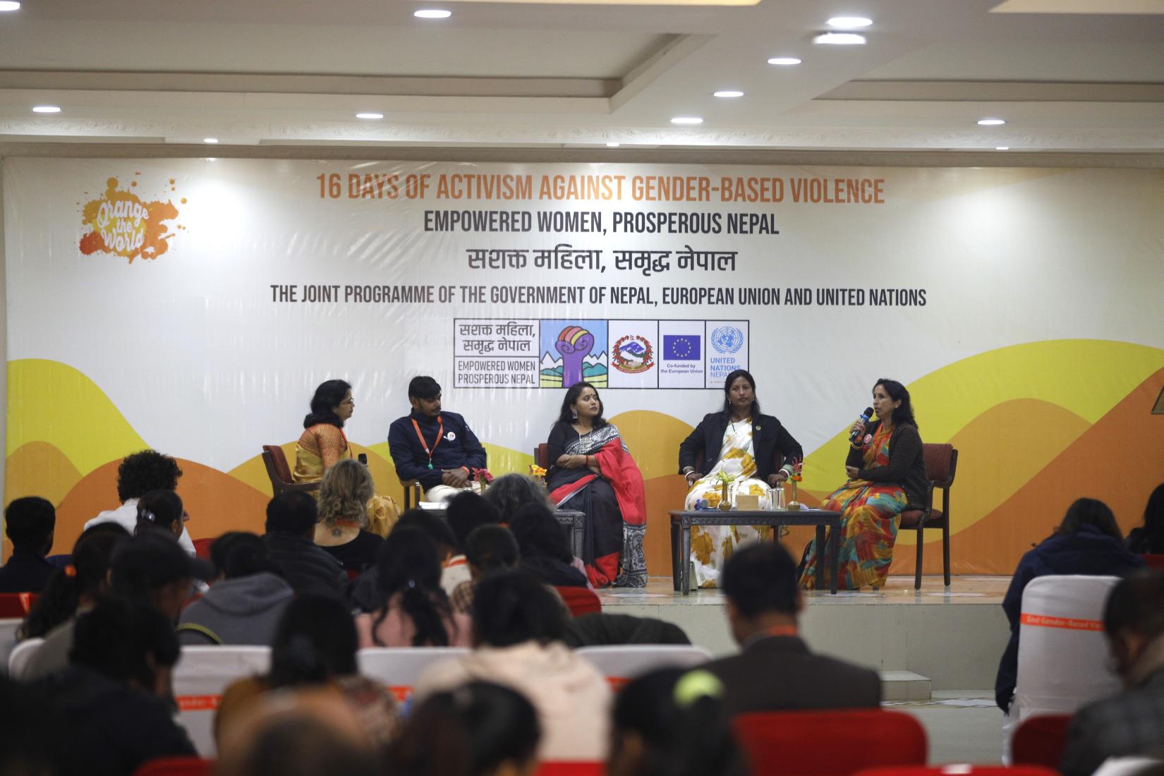 Four women and one man are seated on a platform, engaged in a policy dialogue focused on investing in the prevention of Gender-Based Violence. In the foreground, several individuals are seated, facing the stage, attentively listening to the discussion on this important topic.