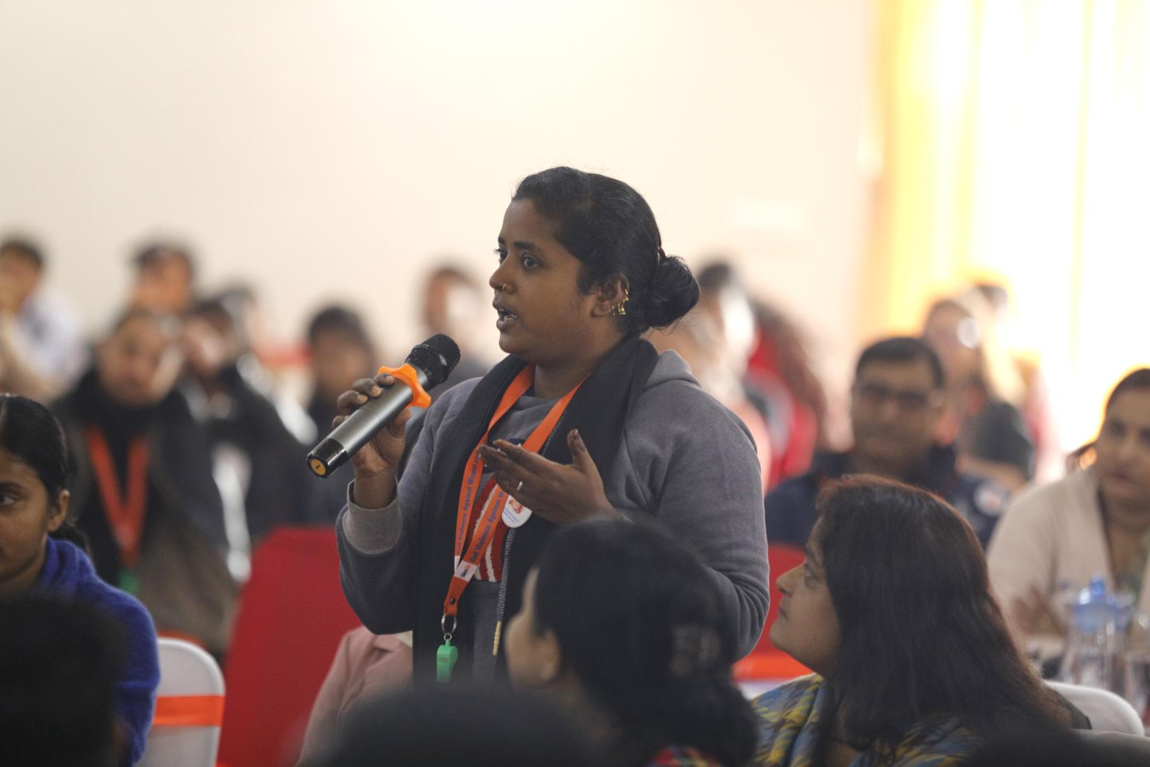 A young girl holds a microphone and speaks while being surrounded by a group of young individuals seated on chairs, attentively listening to her.