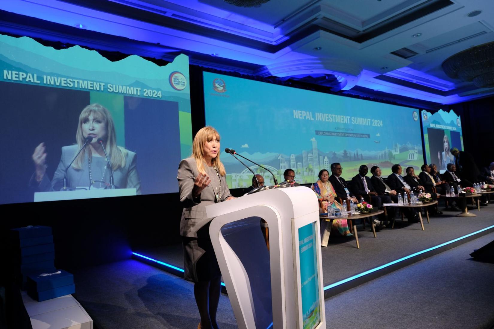 A woman speaking at the podium against a digital backdrop where the text says Nepal Investment Summit.  