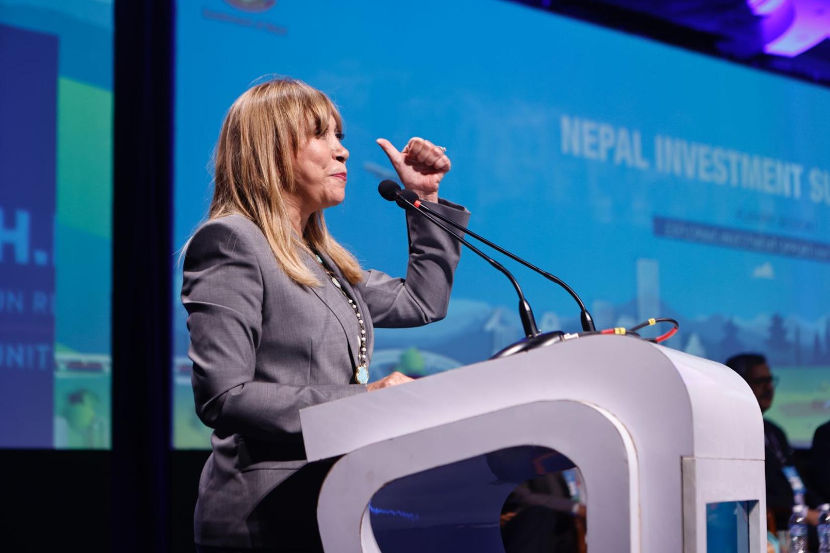 A woman speaking at the podium against a digital backdrop where the text says Nepal Investment Summit.  