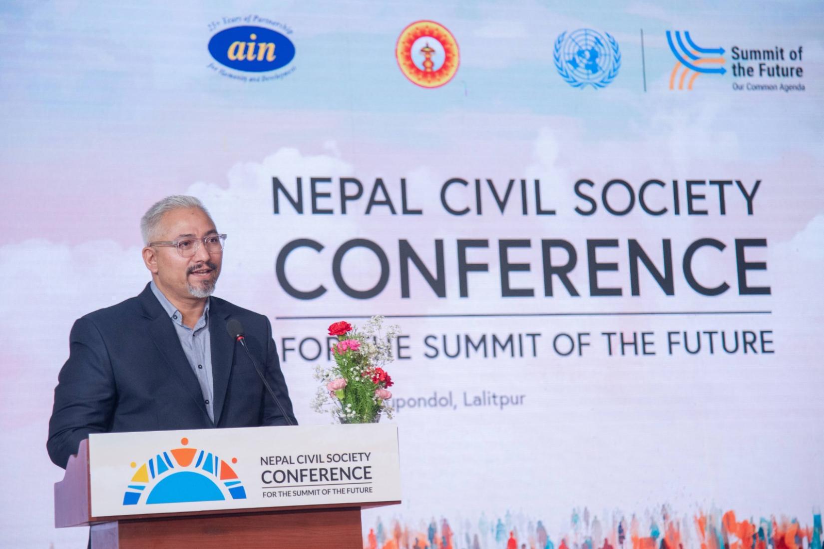 A speaker stands at a podium with a microphone, while the backdrop behind them displays the conference title, " Nepal Civil Society Conference for the Summit of the Future," alongside three logos.