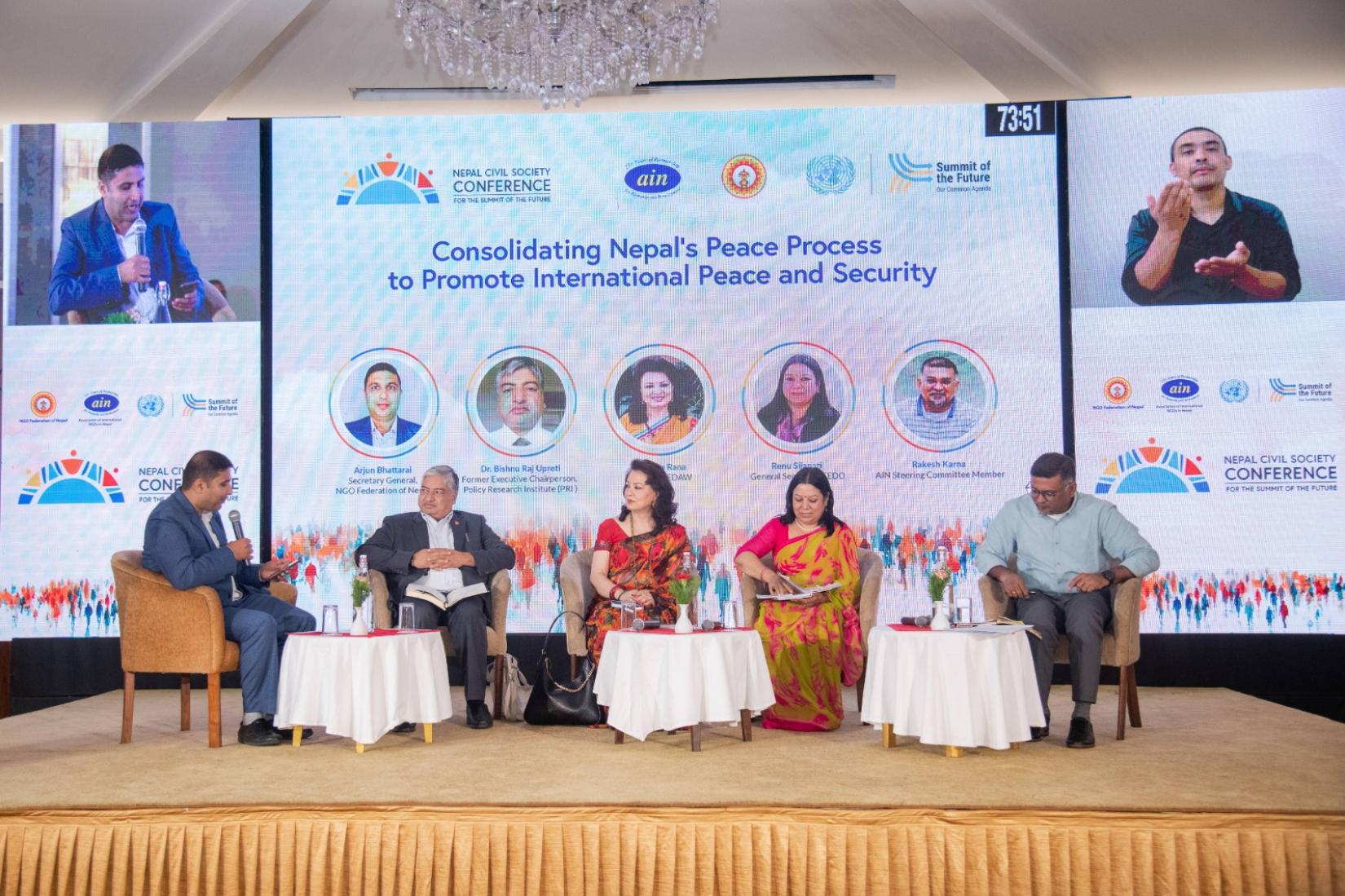 Panel discussion with five speakers on stage, with a banner that reads: Consolidating Nepal’s Peace Process to Promote International Peace and Security in the background and a large screen showing a speaker and a sign language interpreter.