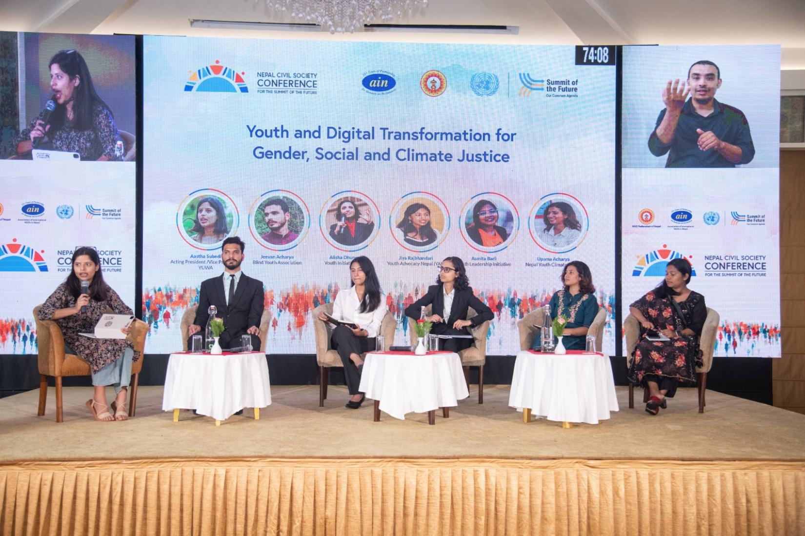 Six people participating in a panel discussion on stage, with a backdrop displaying "Youth and Digital Transformation for Gender, Social, and Climate Justice." The speakers, seated on a stage with a large screen behind them, are engaged in a discussion.