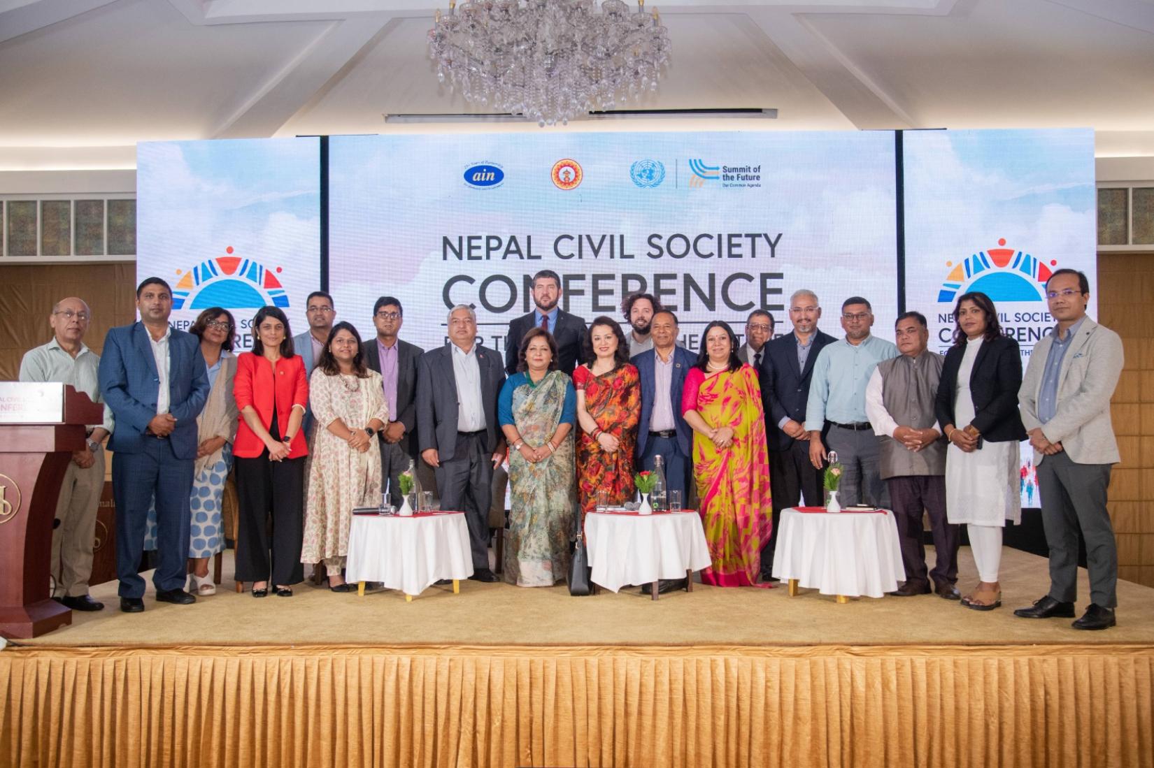A group of 20 people in formal attire pose for a photo on stage under a banner titled: Nepal Civil Society Conference for the Summit of the Future. A podium is visible to the left, with large screens displaying the event logo in the background.