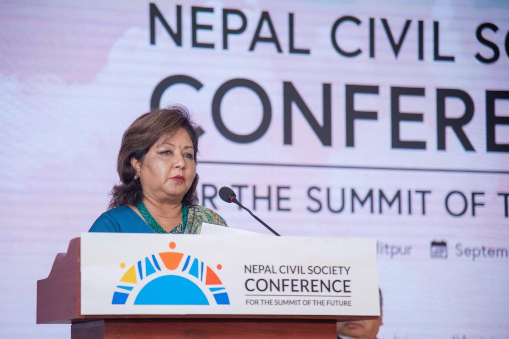 A woman stands at a podium, speaking into a microphone, with a sign behind her displaying the event's name as Nepal Civil Society Conference for the Summit of the Future.