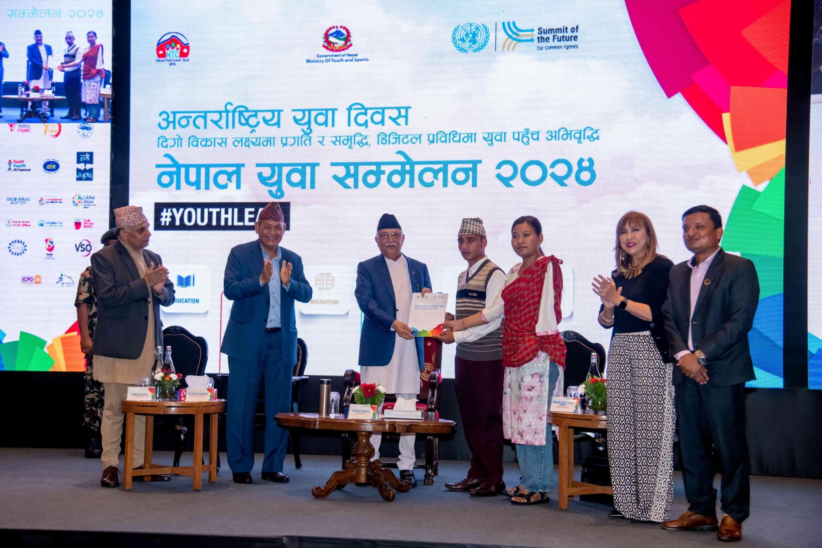 A diverse group of young delegates, consisting of both men and women, stand in a semi-circle proudly displaying the Nepal Youth Declaration document. They are in front of a banner featuring the event's logo and colorful graphics representing youth engagement. The delegates are smiling and wearing badges, indicating their participation in the summit.