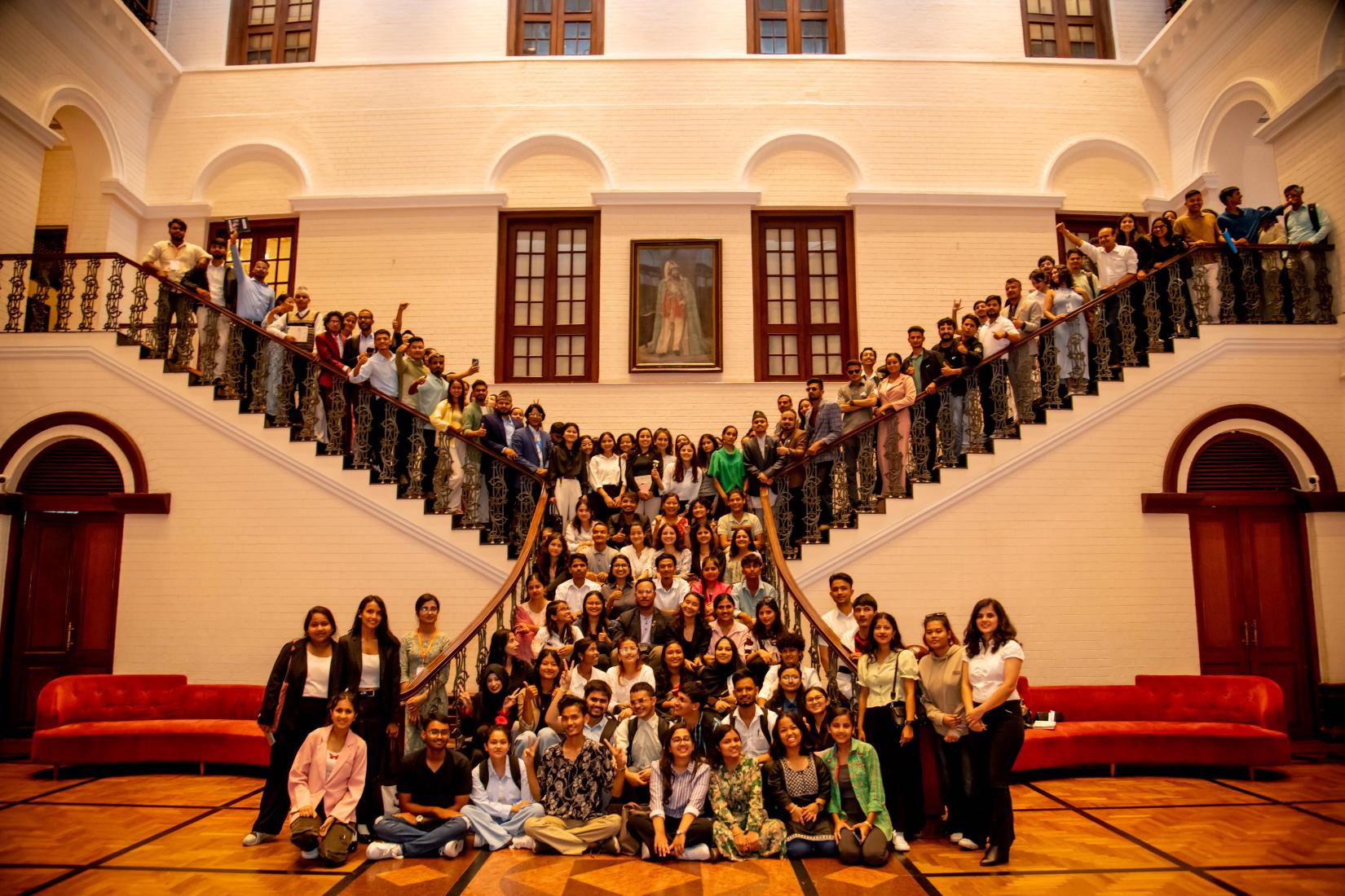 Youth delegates sitting on leader, actively posing for photo during the Nepal Youth Declaration, some of clicking selfie and holding books and a picture at the center. The leader has two ways to go upstairs. Two sofas at ground level.