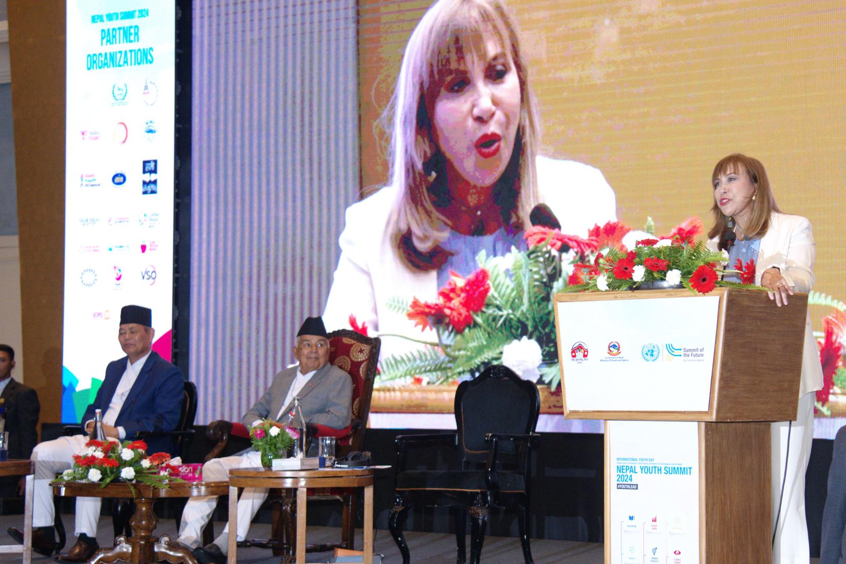 A person with security person behind presents at a podium, while a large screen behind him showcases relevant information for the audience. The other two people are listening.