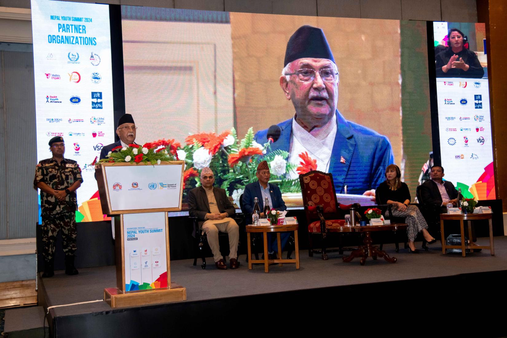 A person with security person behind presents at a podium, while a large screen behind him showcases relevant information for the audience. The other four people are listening.