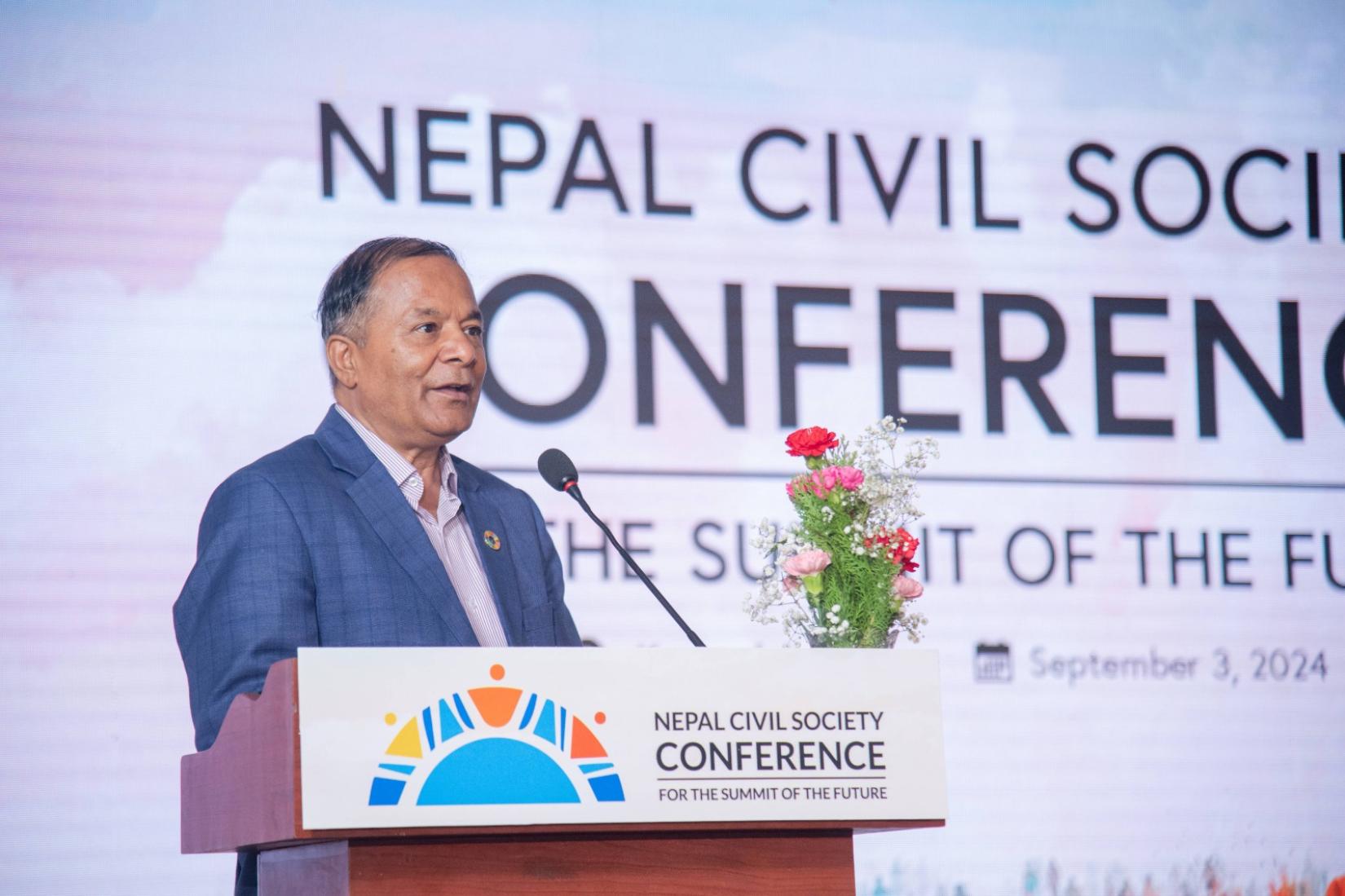 A man in a suit speaks at a podium, with a large screen behind him displaying the conference name Nepal Civil Society Conference for the Summit of the Future and three logos.