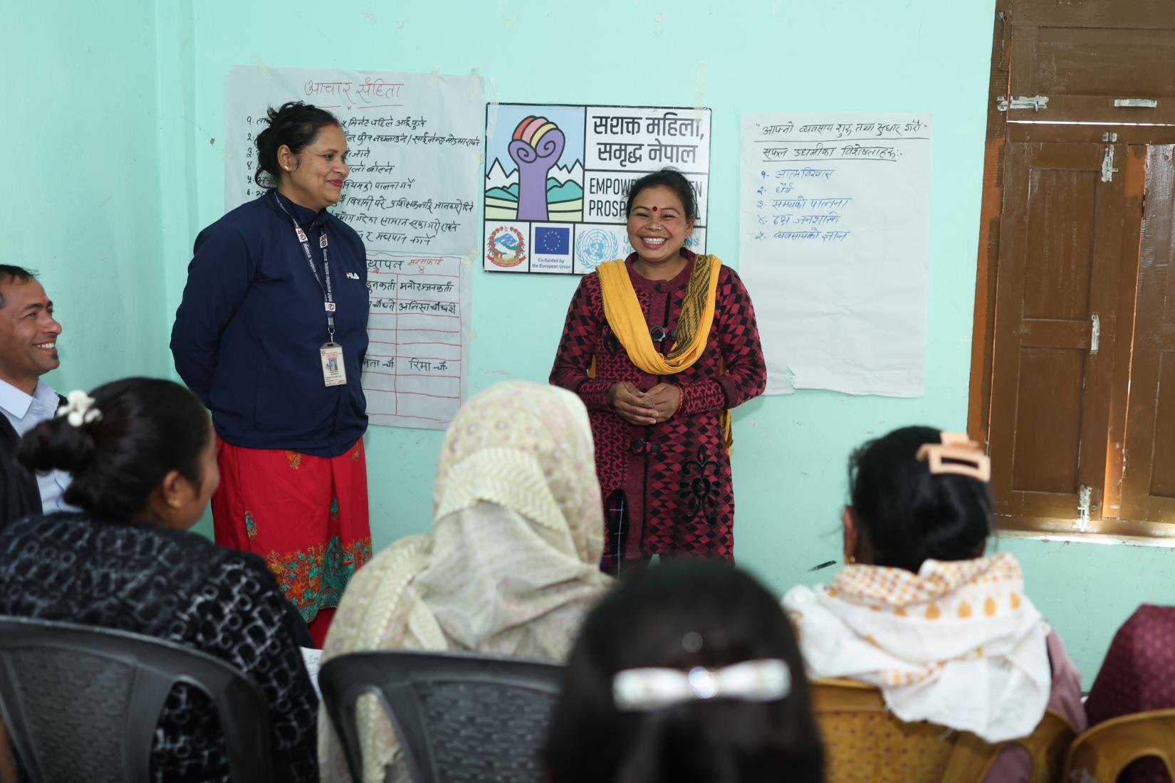 Two women are standing in front of a small group of seated women; one of them is speaking with a warm, smiling expression.