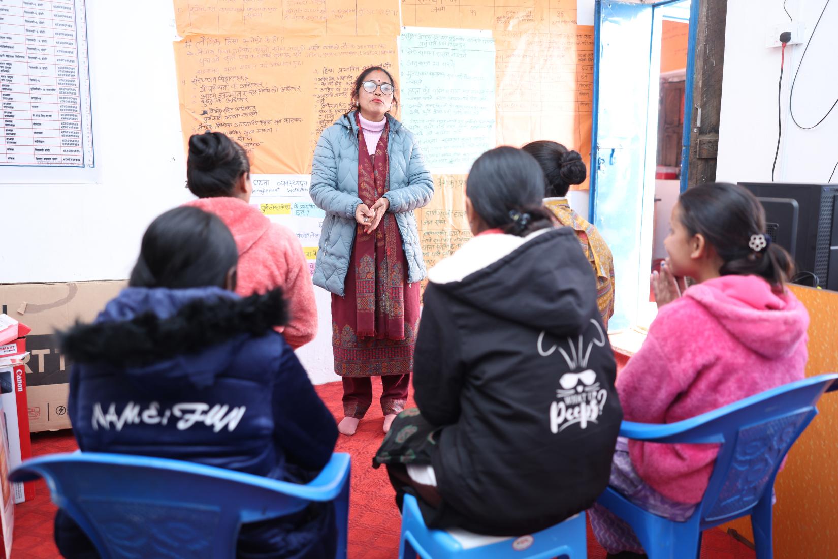One lady is standing while the other is sitting in a chair, facing her and listening attentively.