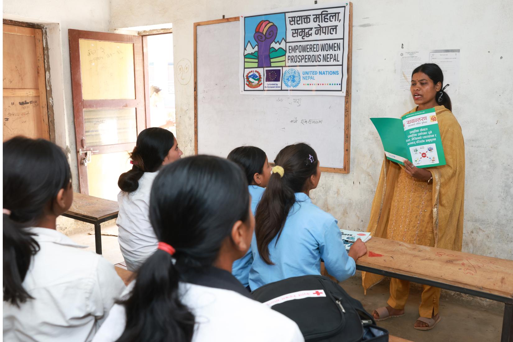 A woman stands at the front of a classroom holding a book titled Rupantaran, while the other students sit on benches, attentively listening to her.