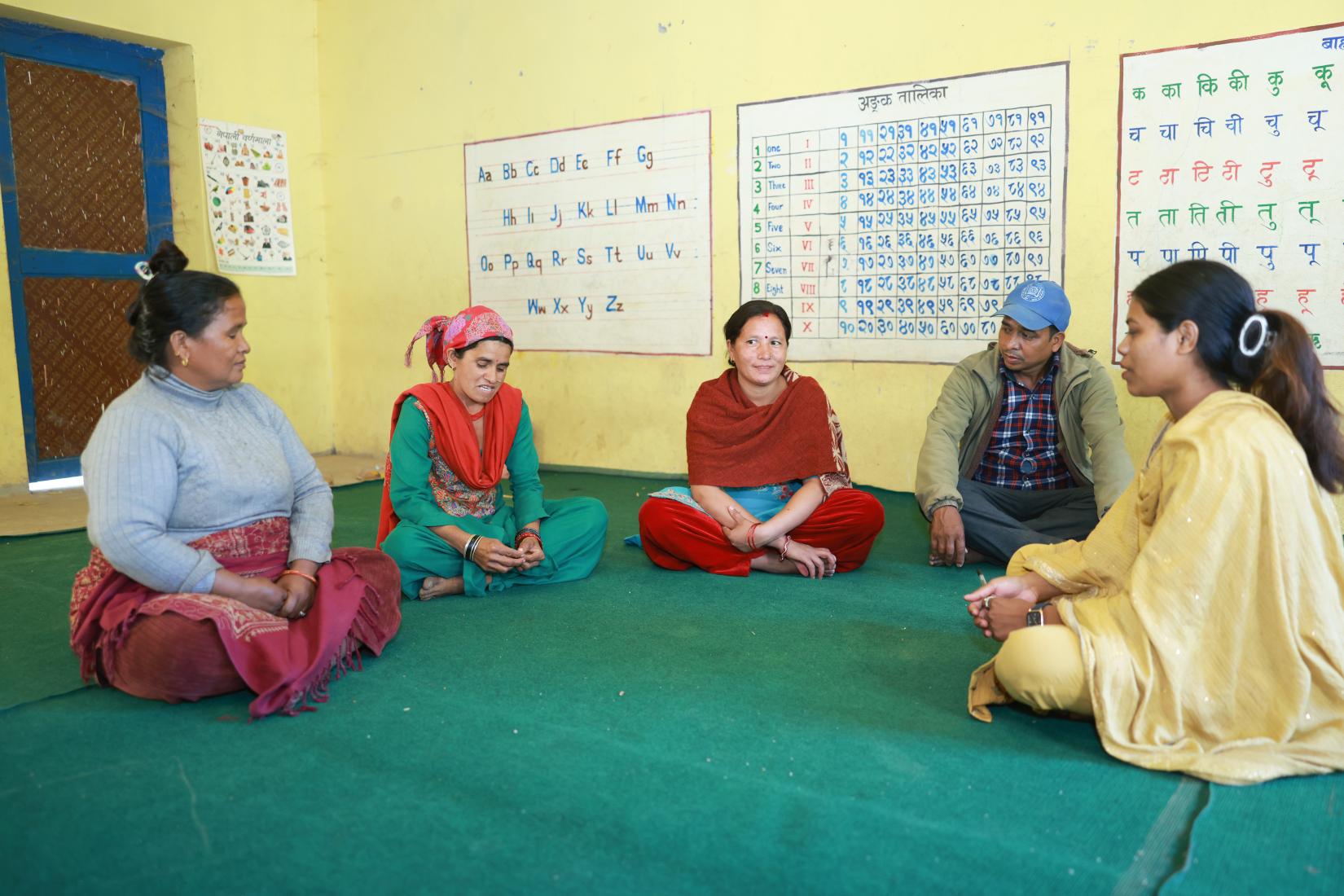 Four women and one man are seated in a room, engaged in a discussion. Alphabet posters are visible in the background.