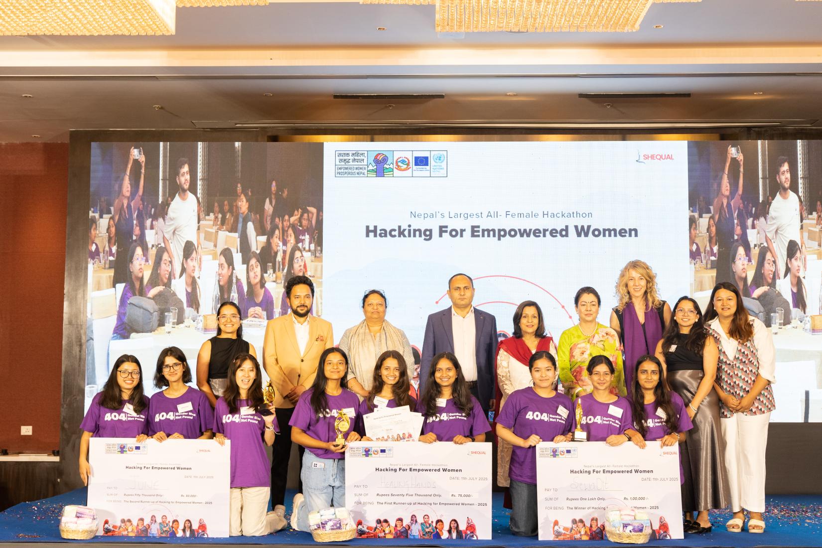 Group photo featuring nine young women kneeling in front, holding a symbolic cheque awarded as a competition prize, with nine individuals standing behind them on stage. A large banner in the background displays the hackathon theme: 'Hacking for Empowered Women'.