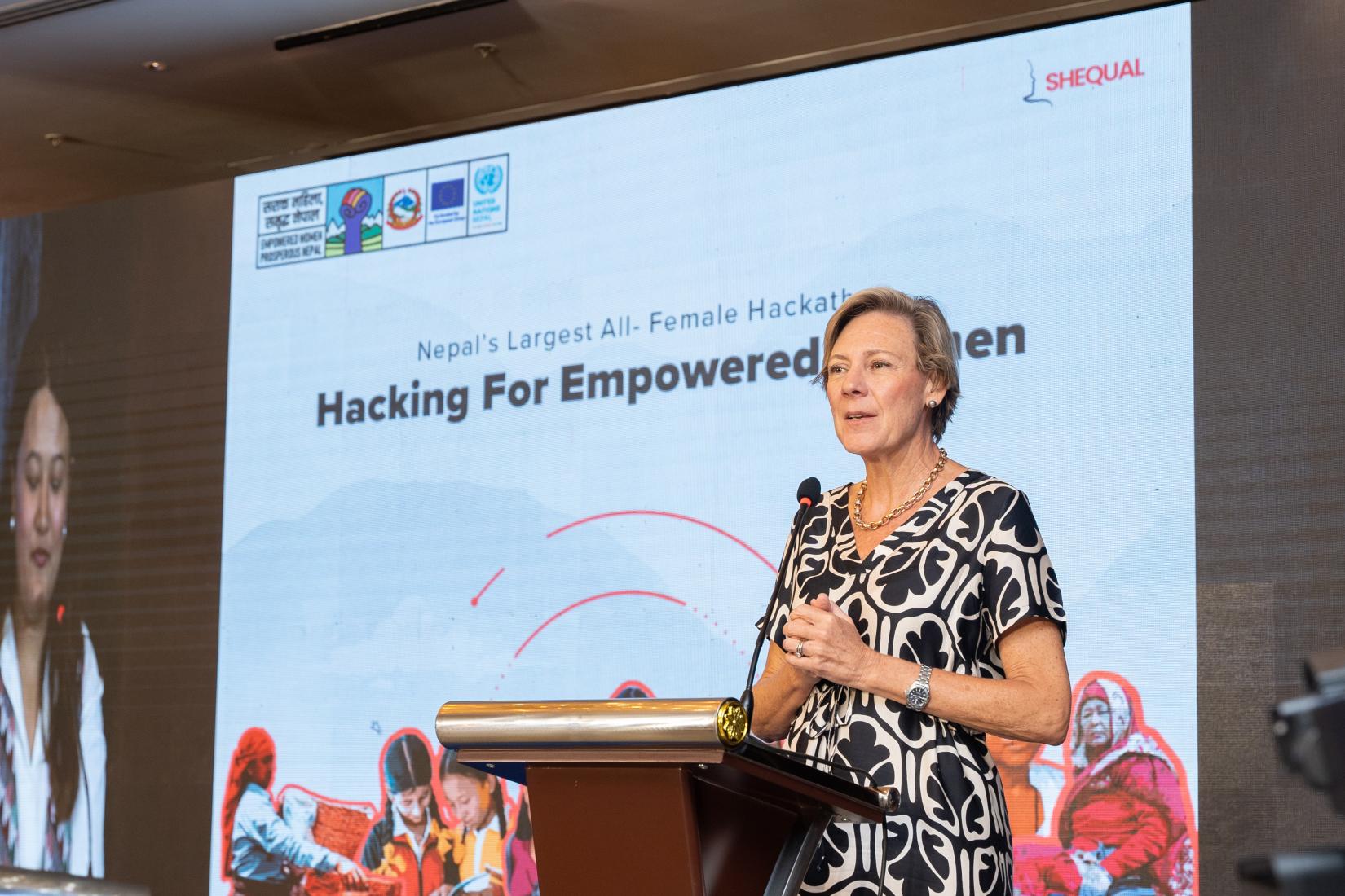 A woman standing at a podium delivering a speech, with a banner in the background that reads 'Hacking for Empowered Women'.
