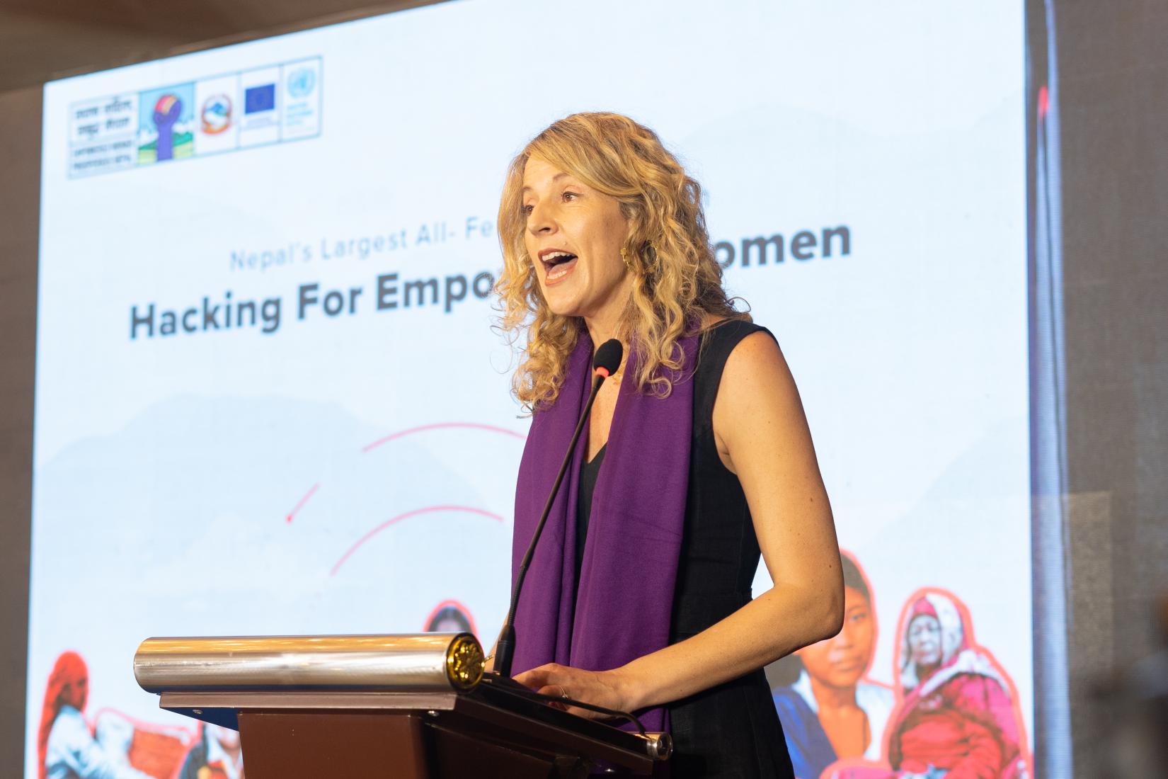 A woman stands at a podium delivering a speech, with a banner behind her that reads 'Hacking for Empowered Women'.
