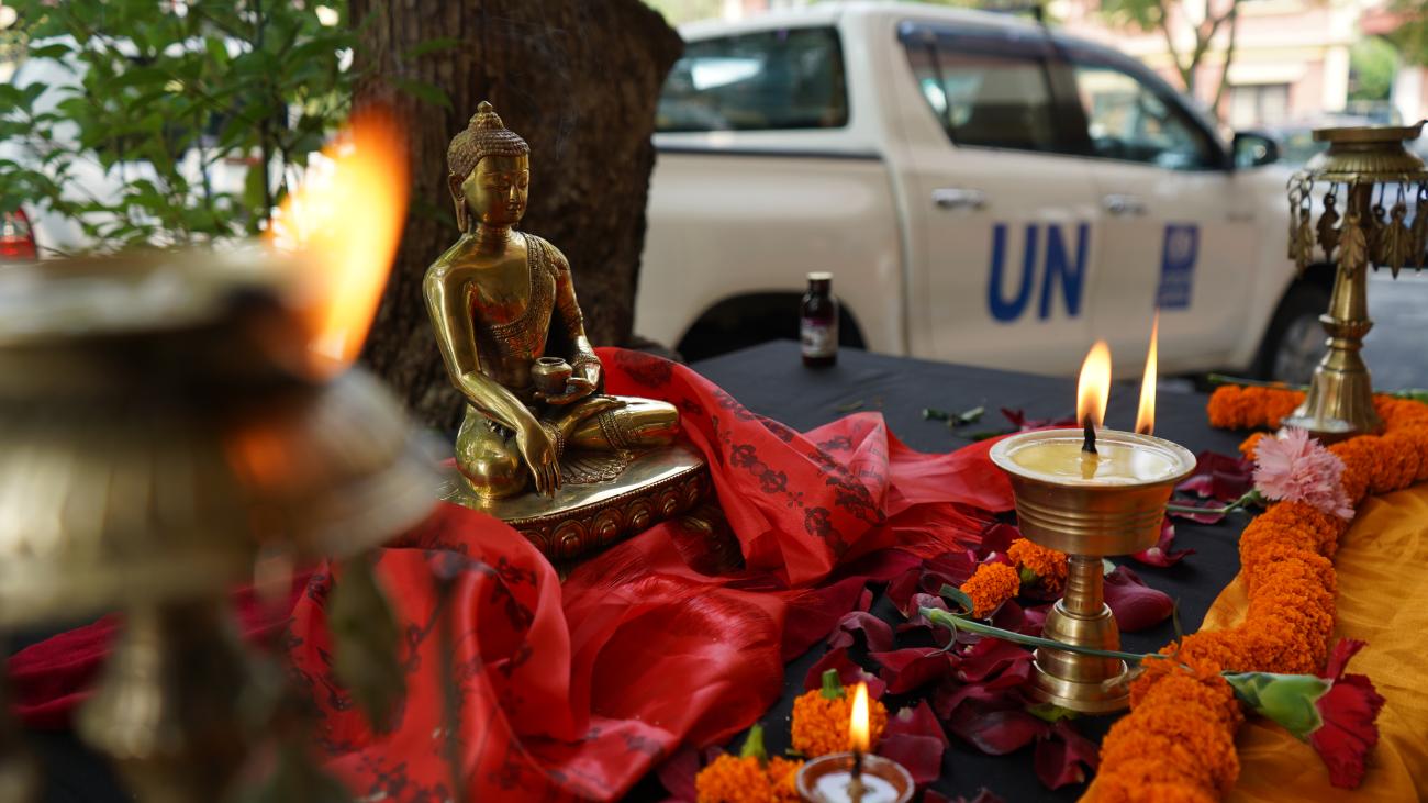 Golden Buddha surrounded by candles, flowers and red silk shawls. Behind the statue is the trunk of a tree and a UN car is visible in the background.