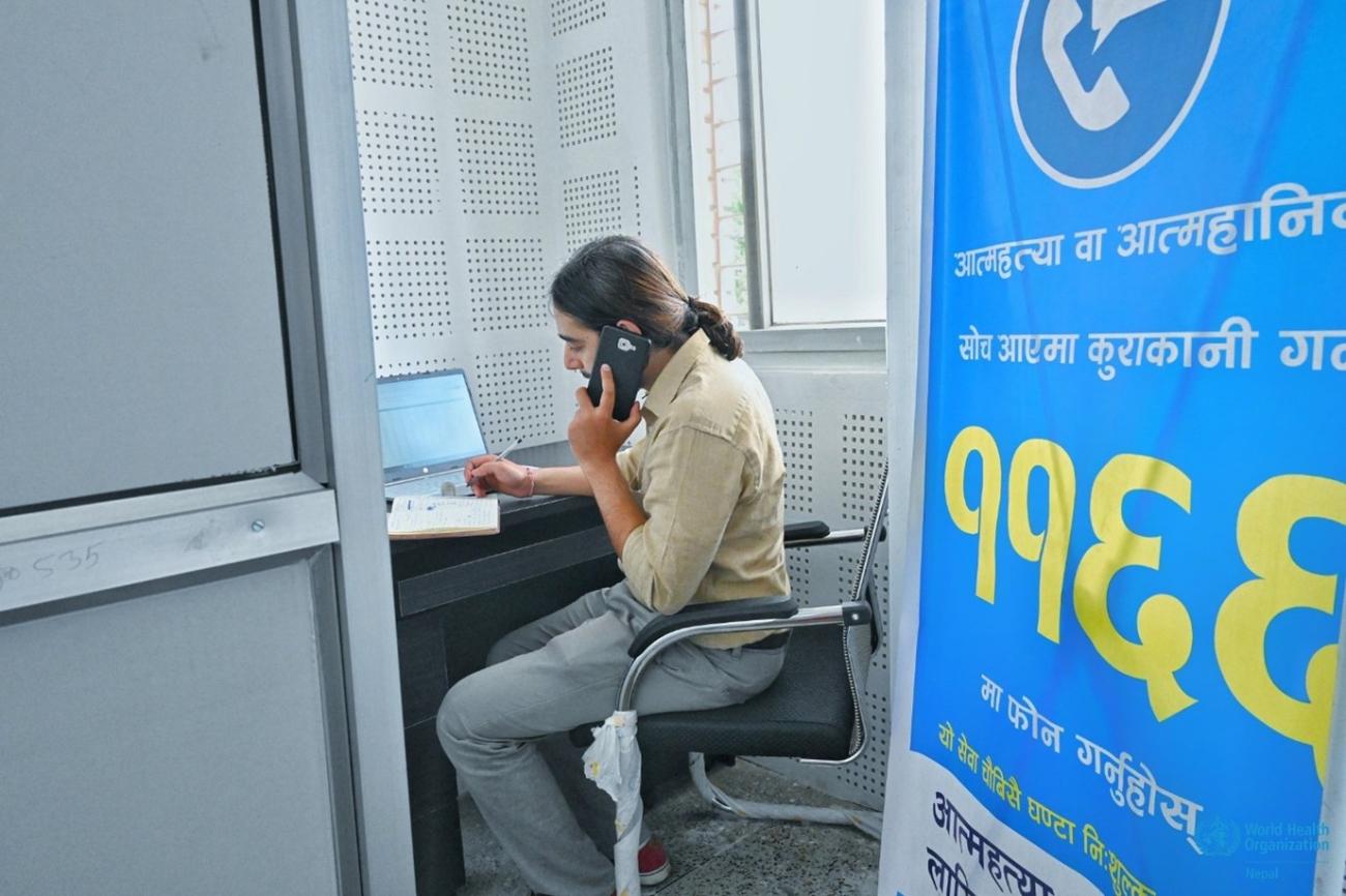A man sits on his desk in front of the laptop inside his cubicle. He is holding his mobile and writing down on his notebook.
