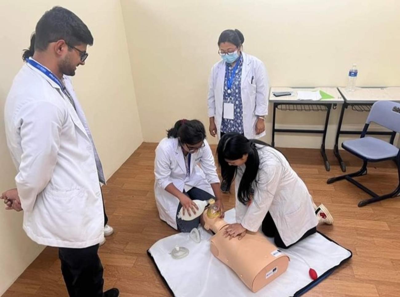 Two women with white coat practicing CPR on a dummy on the floor while a man and a third woman with white coat watch them inside a room.