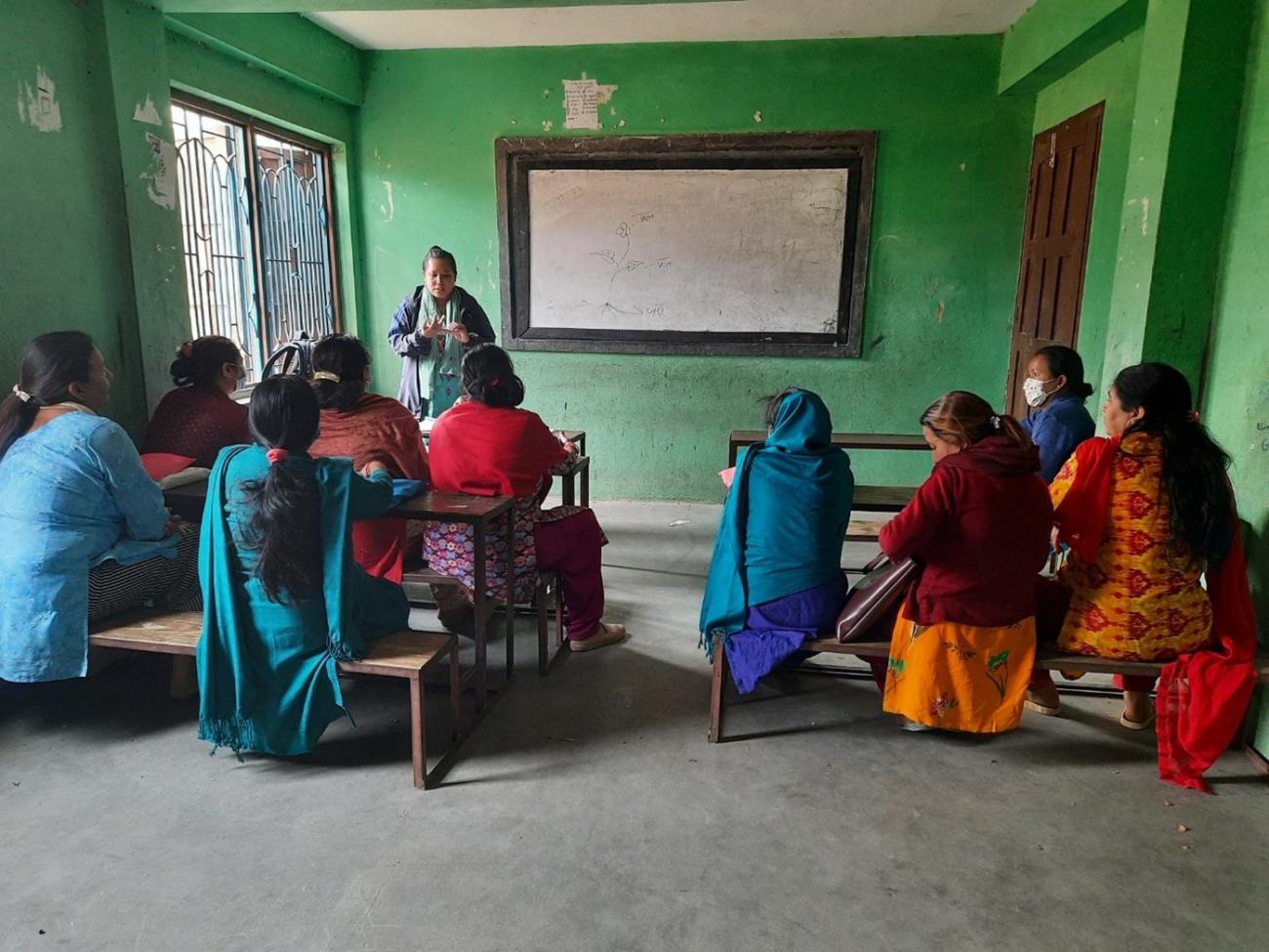 n a classroom setting, the mental health activist is standing and explaining to all the women sitting on benches with their backs faced to the camera. There are around nine participants listening to the activist.   mental health activist discusses with female community health volunteers on early identification of people in distress, 2021. 