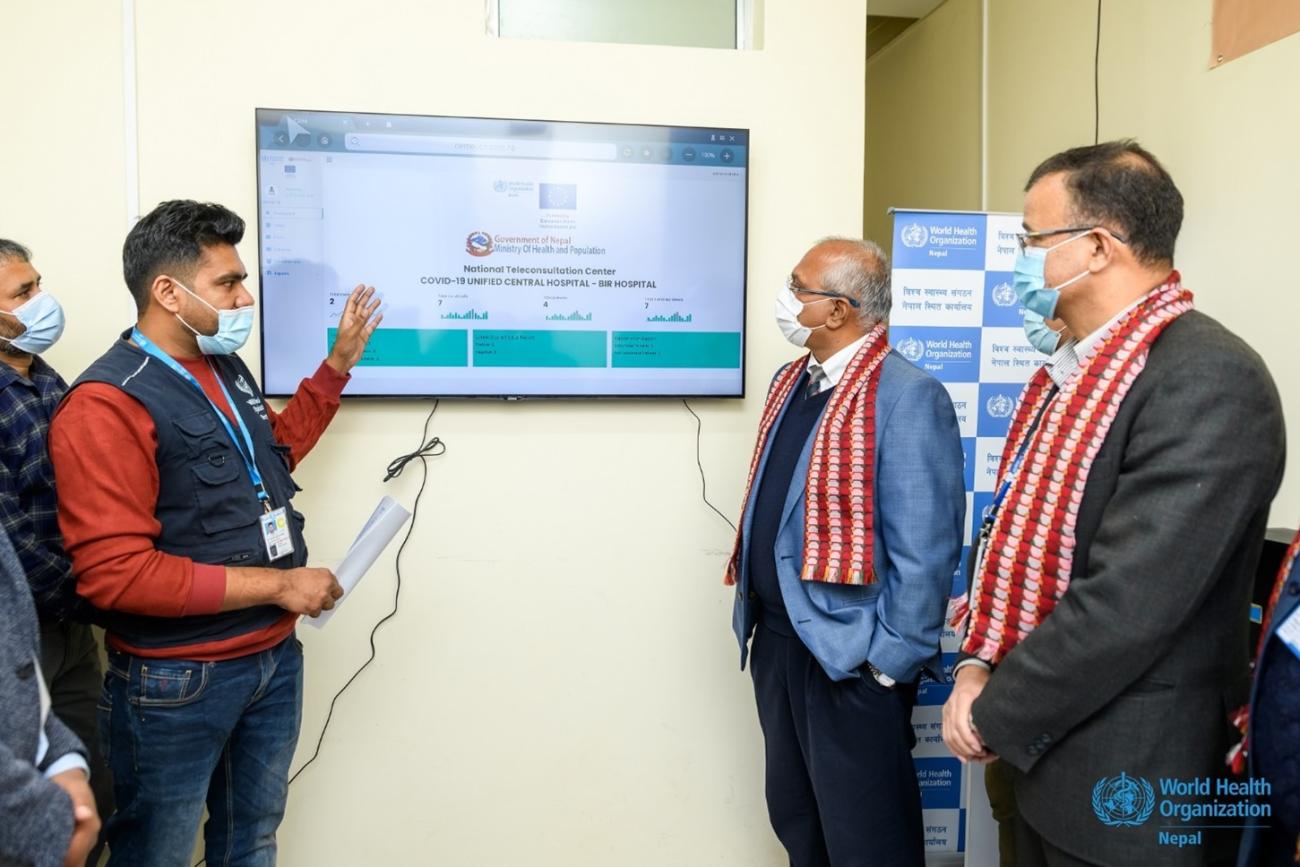 A man points at the tv screen explaining two other men standing in front of him at the teleconsultation center.