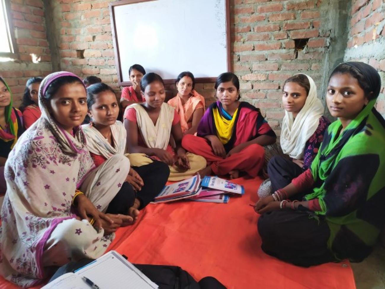 A group of eleven women wearing Kurthas are sitting on a red mattress inside a newly built room. There are notebooks in front of them and a whiteboard behind at the wall.