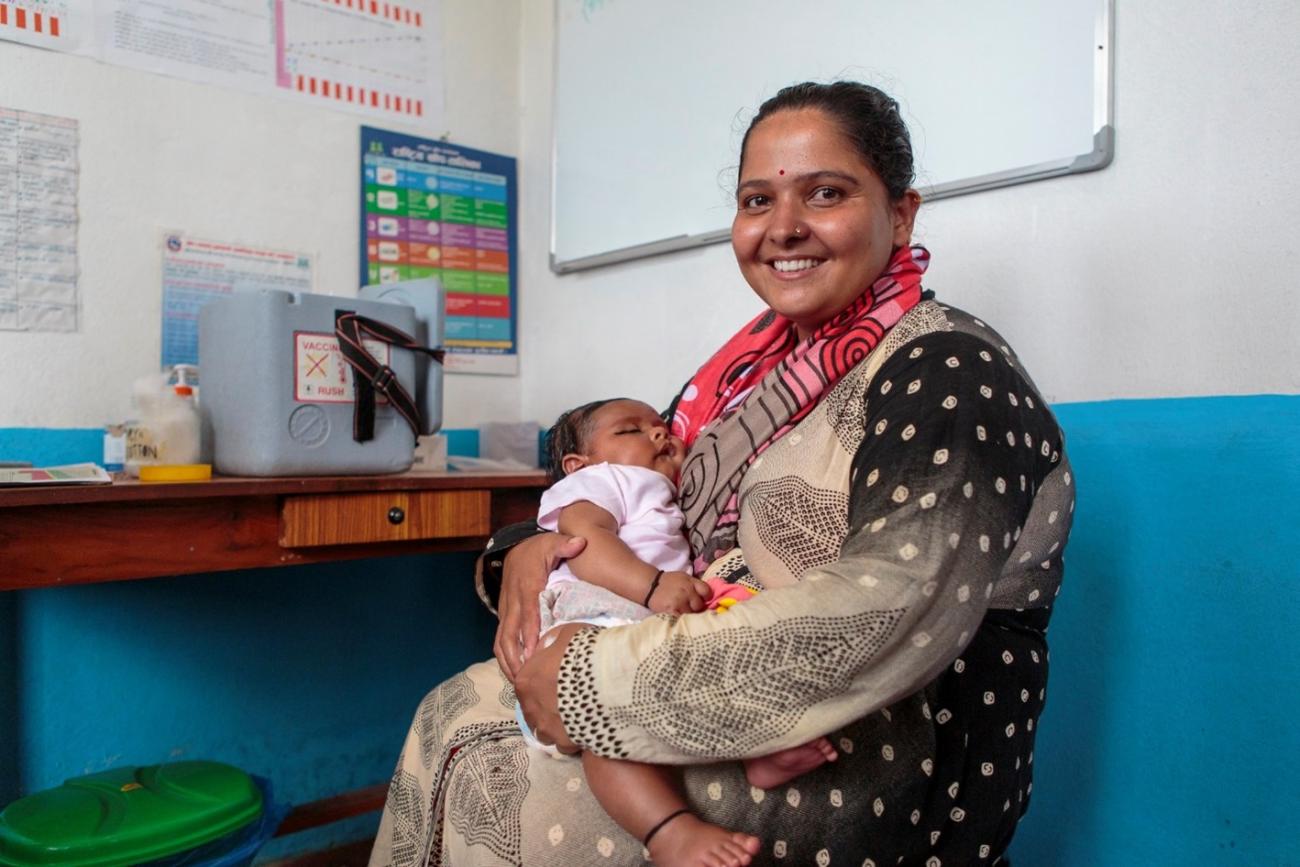 A mother holds her sleeping baby who's a few months old with a big smile on her face. The woman is sitting on a chair inside a room at the health post.