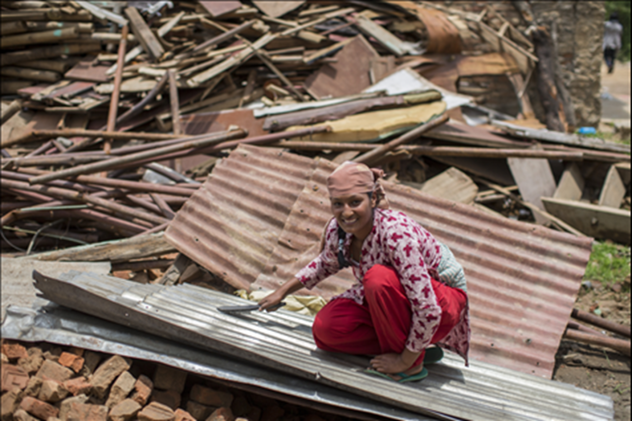 A woman wearing red kurtha and a scarf tied on her head looks at the camera with a smile as she examines the tin roof to rebuild a house.