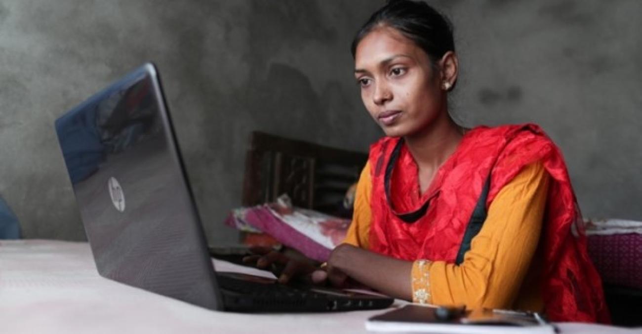  A woman wearing yellow Kurtha and red shawl is using a computer with a notebook beside it.