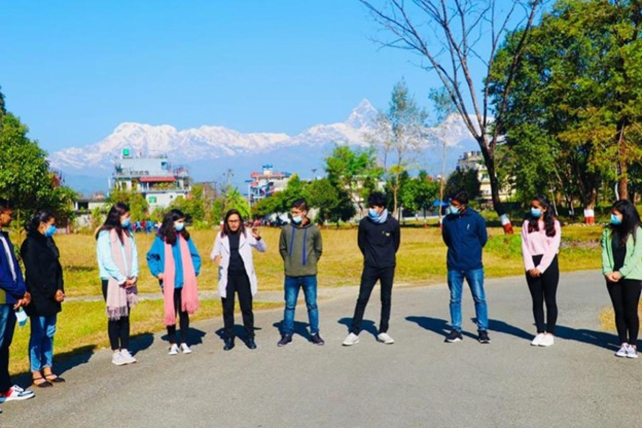 10 people stading on the ground in a line while a woman in the middle has her hand raised, saying something to the rest of the people. The background has beautiful mountains.