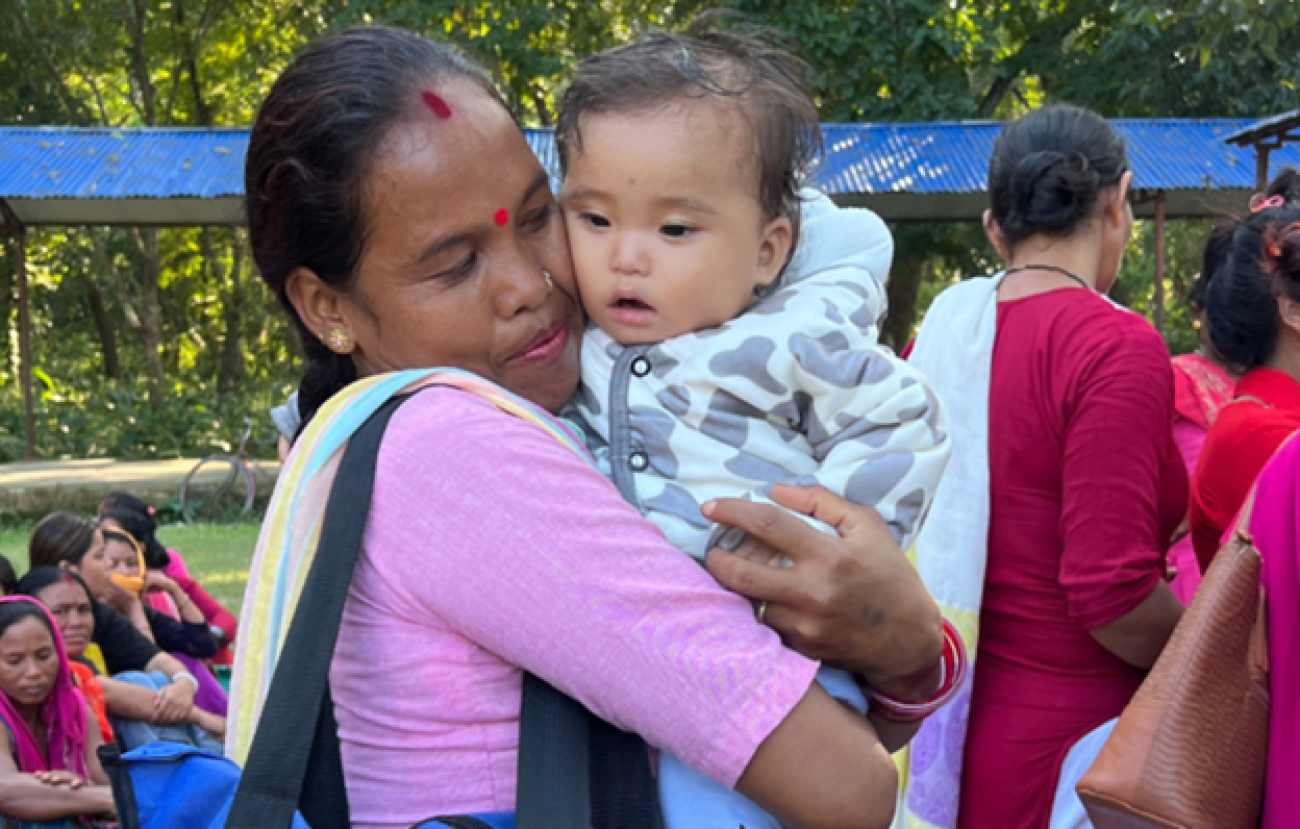 A woman in a pink sari and a colorful scarf hugs a baby wearing a light blue cow-print puffer jacket. In the background there is a blue tin shed and other women.