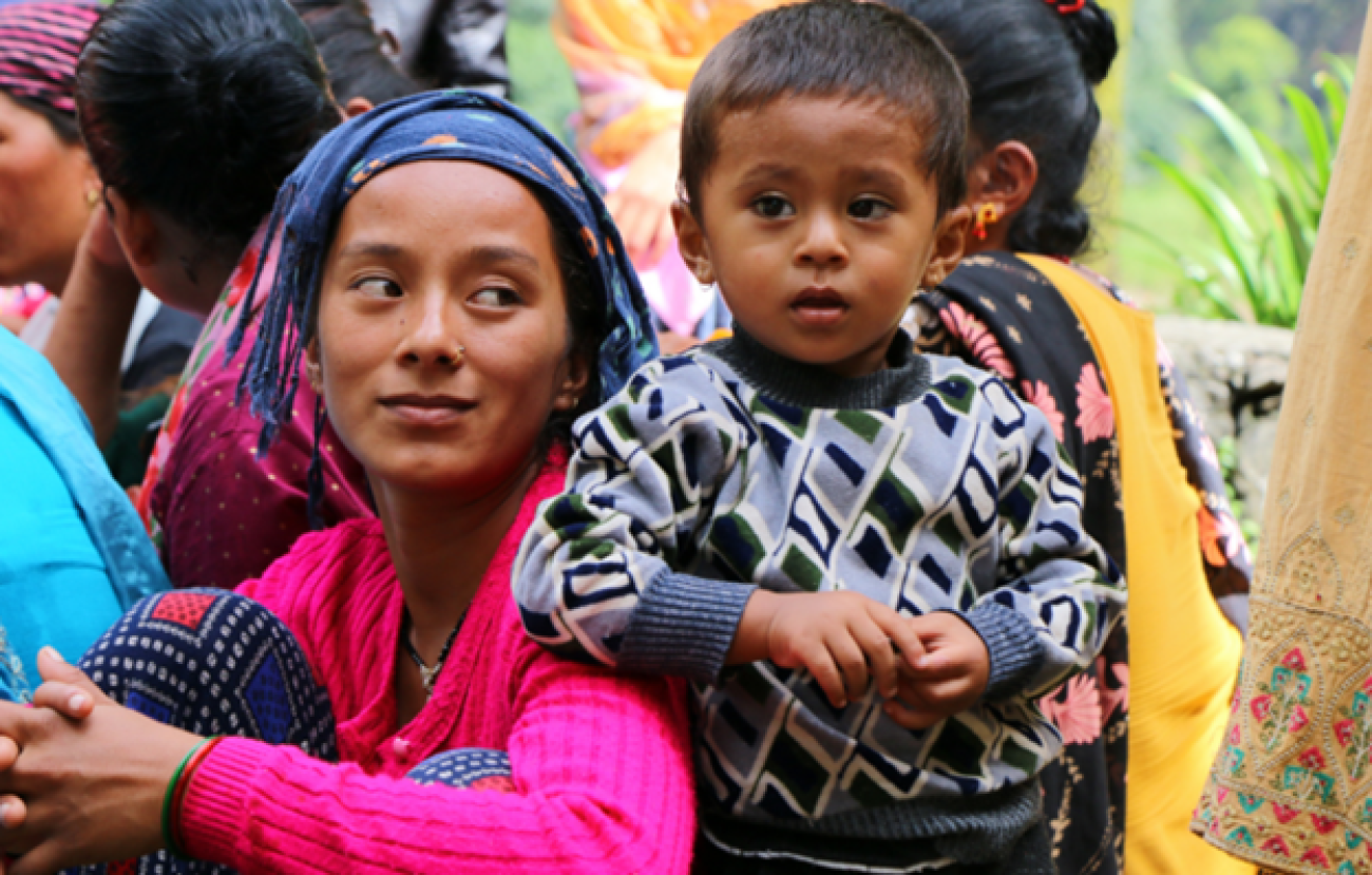Woman in a pink sweater and a navy blue headscarf looks at a little boy who stands next to her gazing into the distance.