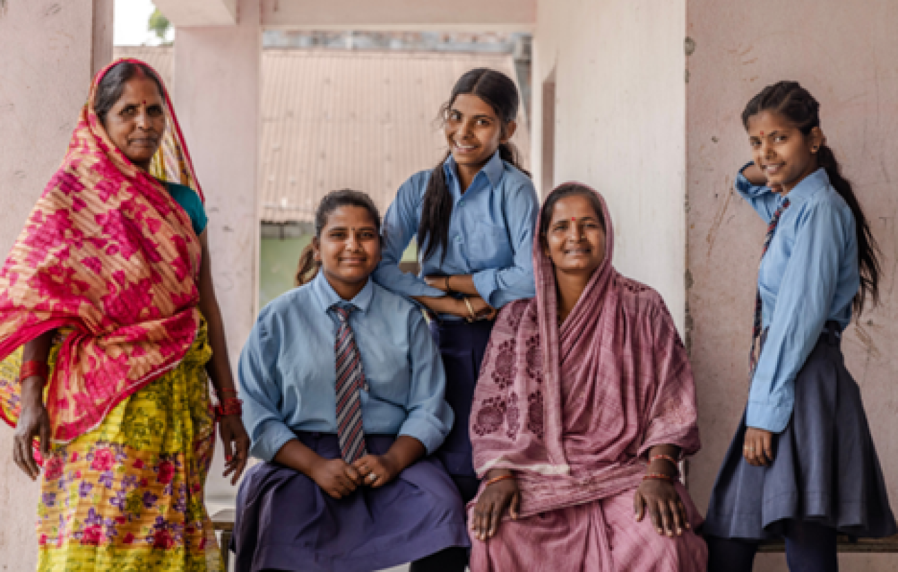 Three school girls wearing their school uniform blue shirt and blue skirt, are looking forward and smiling. There are two other middle aged women wearing sarees and smiling too.