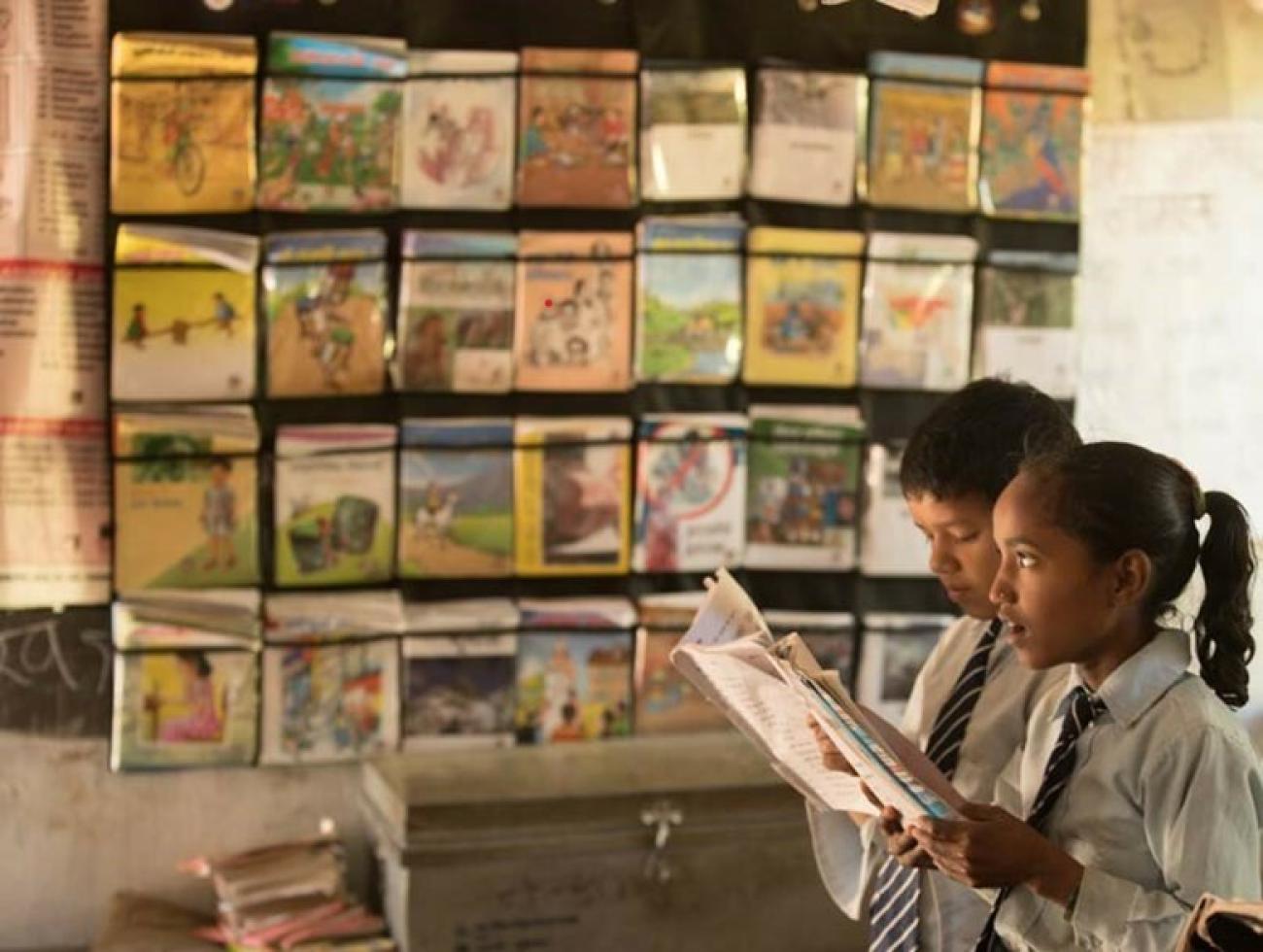 A Nepali girl and boy wearing their white school uniform and striped tie are holding a notebook and reading it in a magazine store