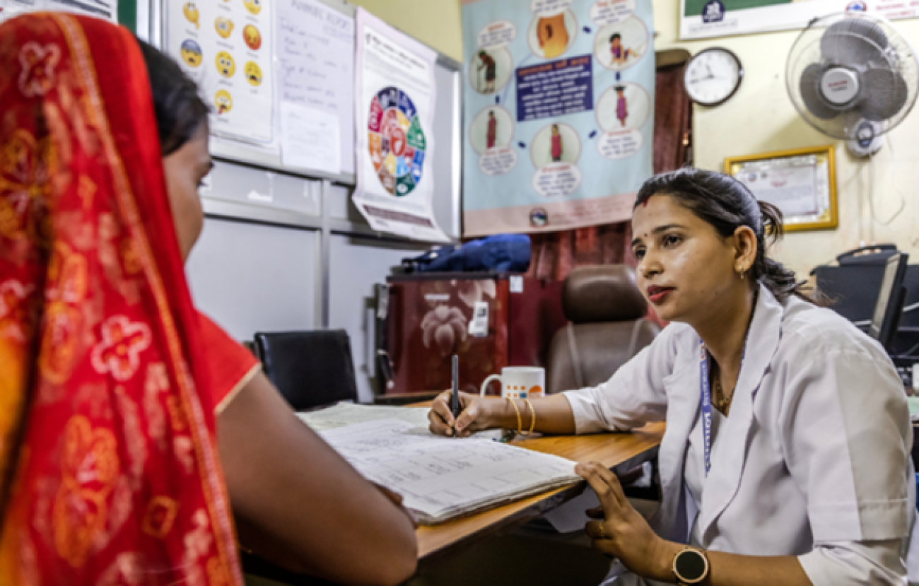 Psychosocial worker Kesh is wearing a white coat and intently listening to the woman in front of her wearing a red saree inside her office.
