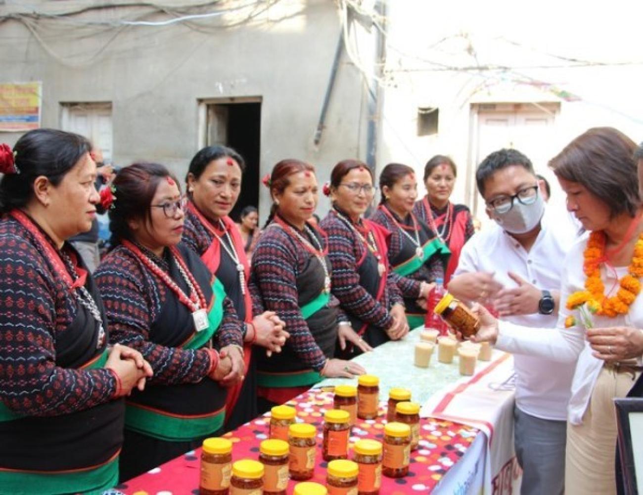 Seven women wearing traditional Newari saree is standing behind a table full of pickle jars and selling them to customers.