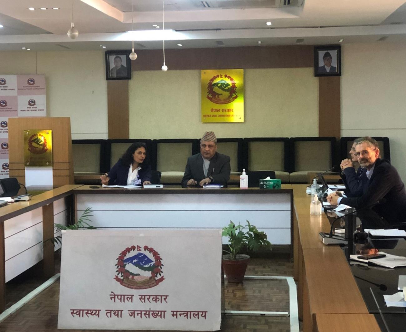 A woman talks to a man sitting beside her in the conference room. There are two more men at the right side, and a board in the middle with the government logo on it.