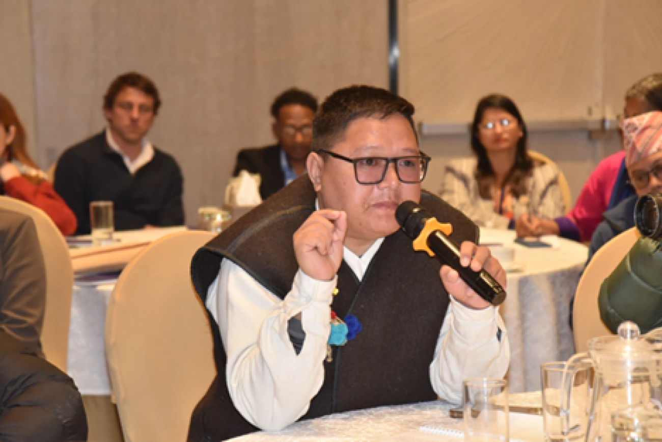 A man in a brown coat and glasses sits amongst others one of many tables and speaks into a microphone.