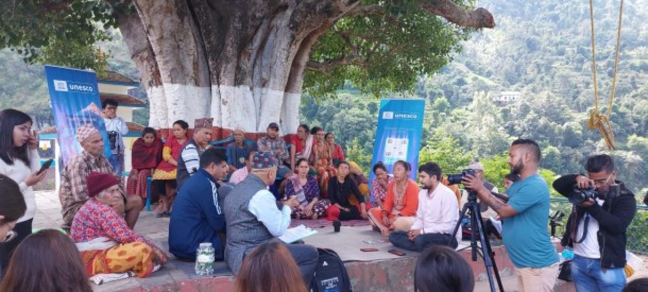 A group of locals sit on a seating area around a tree whilst journalists take pictures. Two blue UNESCO banners are positioned amongst them with trees in the background.