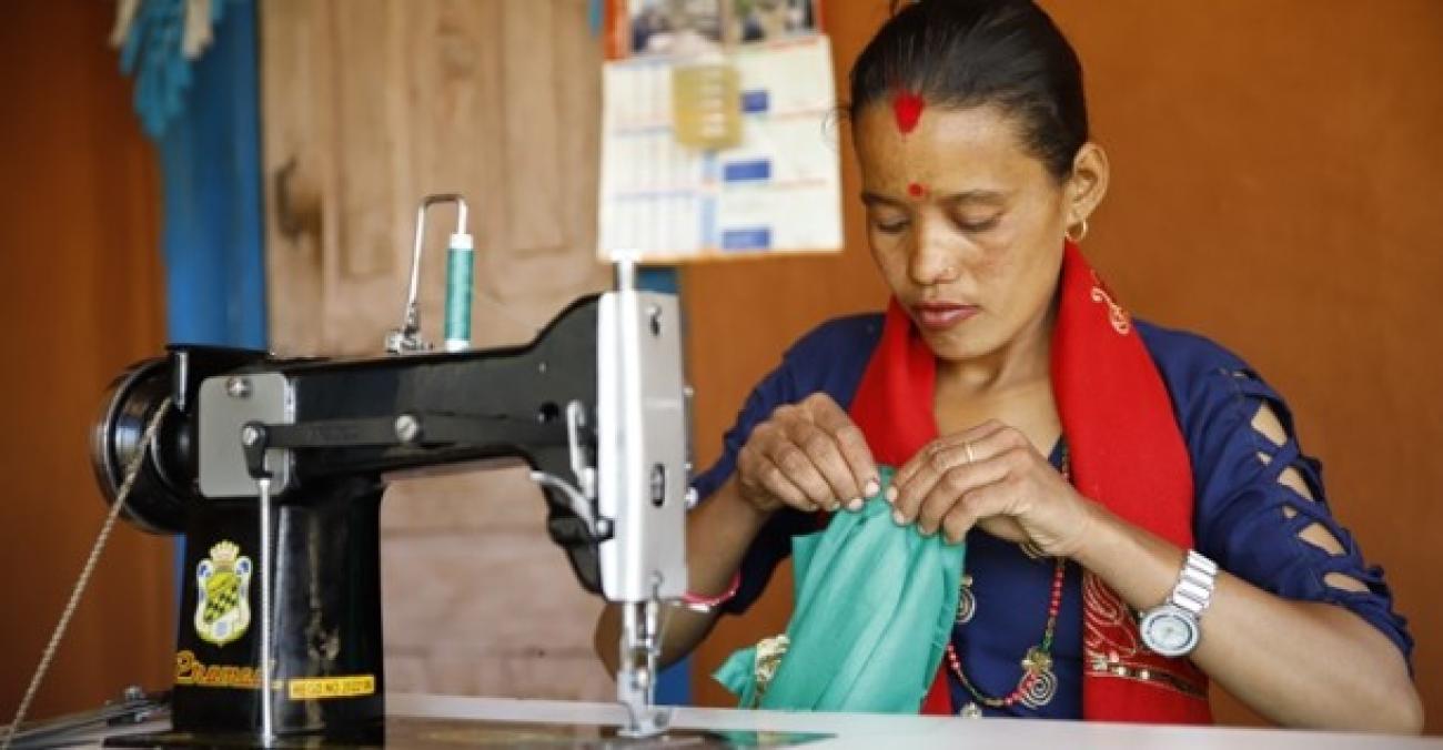 Harikala holds a blue cloth that she is stitching with the black sowing machine in front of her.