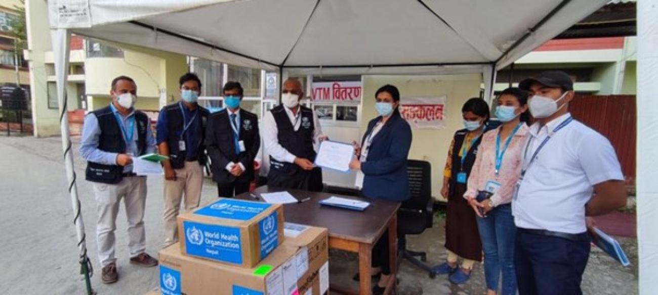 WHO country officer handing a certificate to the woman beside him, while the other six people are standing in the sides inside a tent at the compound. There are boxes with WHO logo at the very front. 