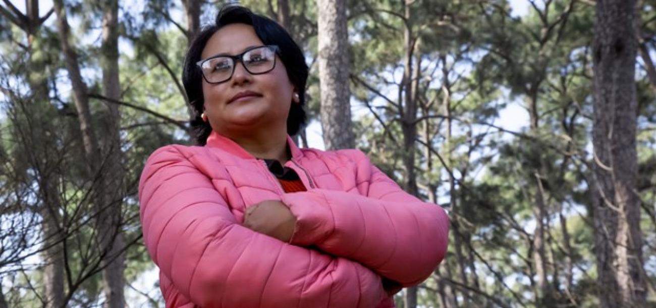A women in a pink jacket and glasses, stands in the woods with her arms crisscrossed gazing into the distance.