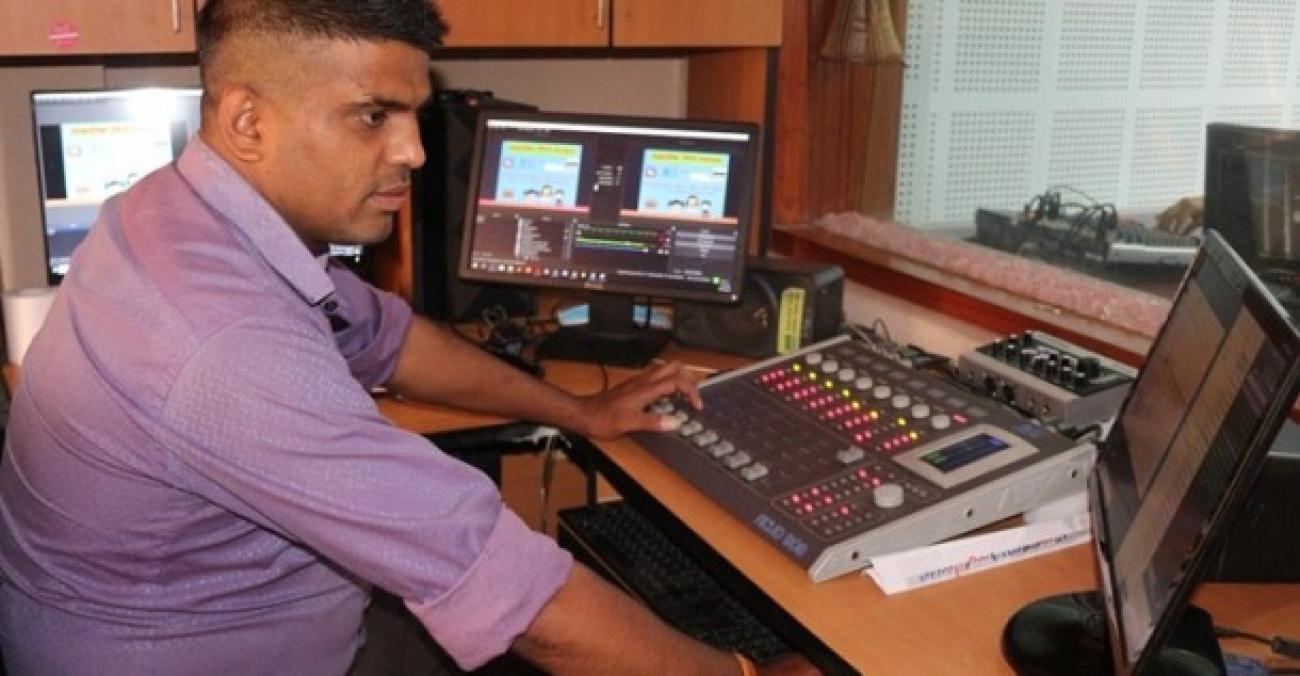 A person wearing purple shirt sitting at a desk using a computer, with two other computers at his left side.