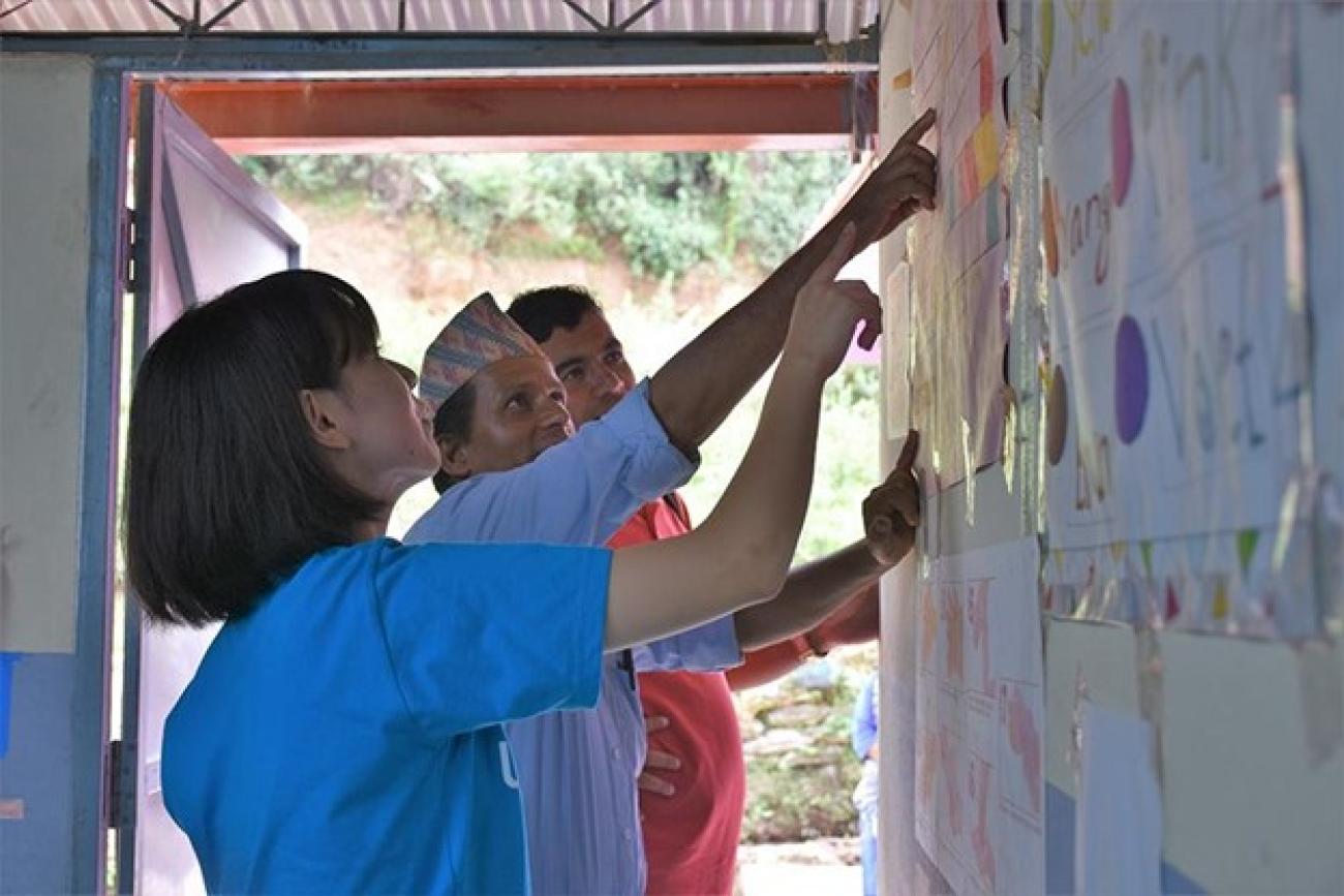 A woman in a in a blue T-shirt stands next to two men: one in a blue shirt and a dhaka topi, the other in a red shirt. The all point to the colorful posters on the walls.
