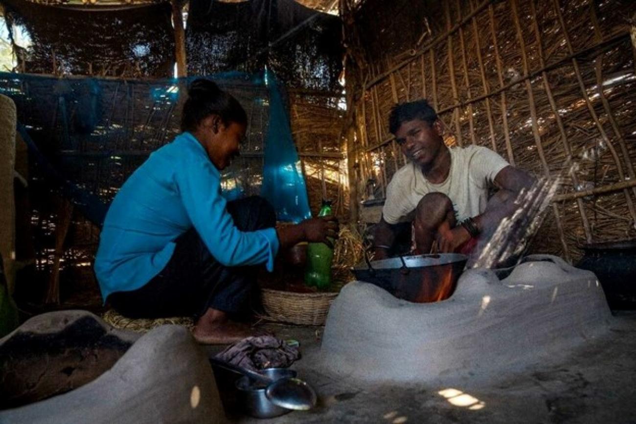 A man in a white T-shirt and a woman in a blue shirt with a green plastic bottle in hand, sit crouched in a straw shelter cooking with metal pots.